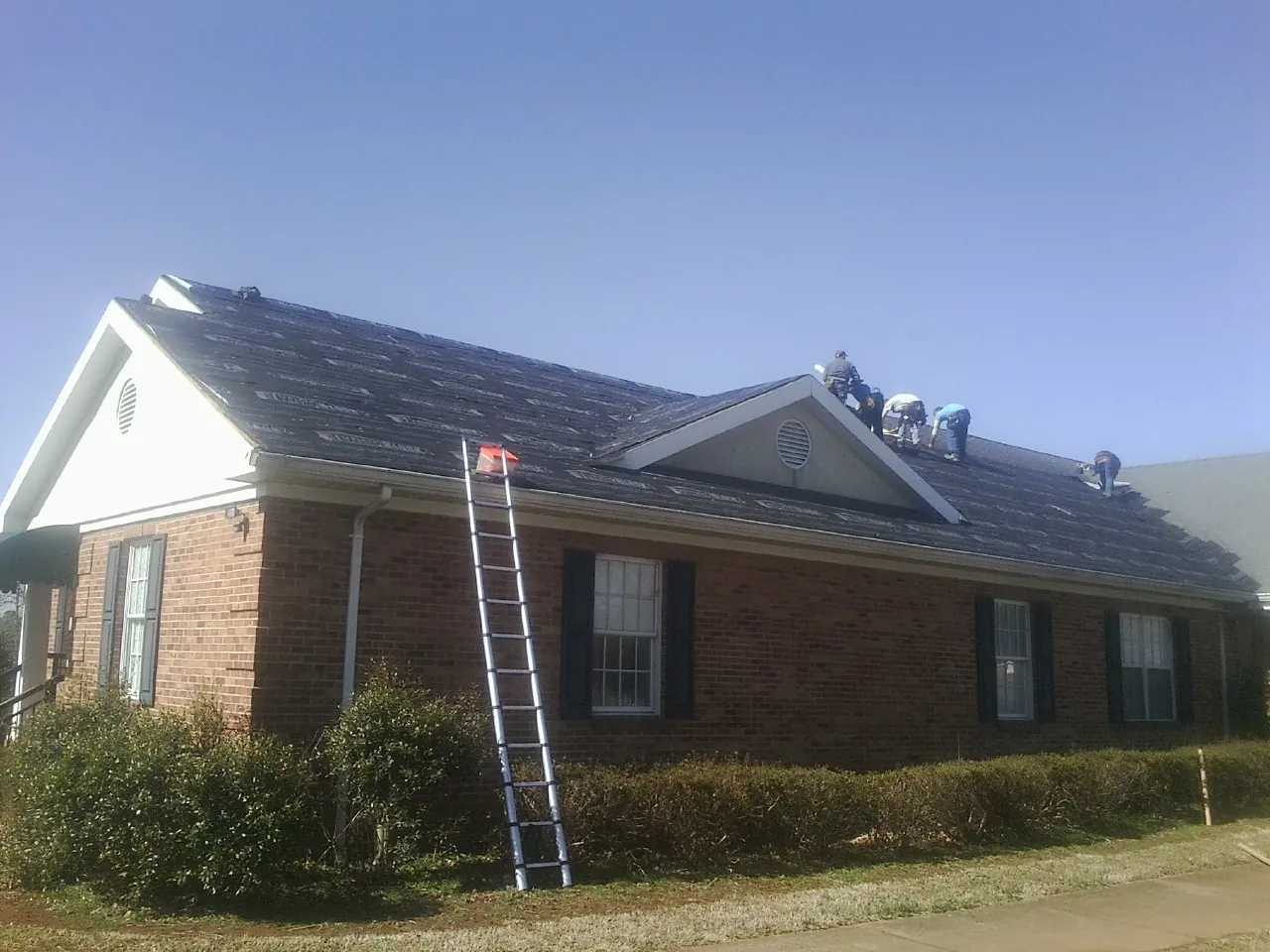 Crew tearing shingles off a brick ranch roof with a ladder set — Emergency Roof Repair work by our crew in Lamy
