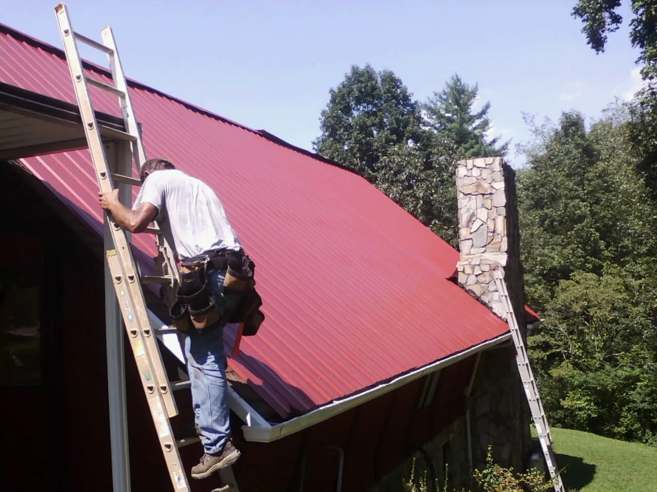 Roofer on a ladder beside a red metal roof and stone chimney — Soffit & Fascia Repair by Roofing Brothers in Lamy